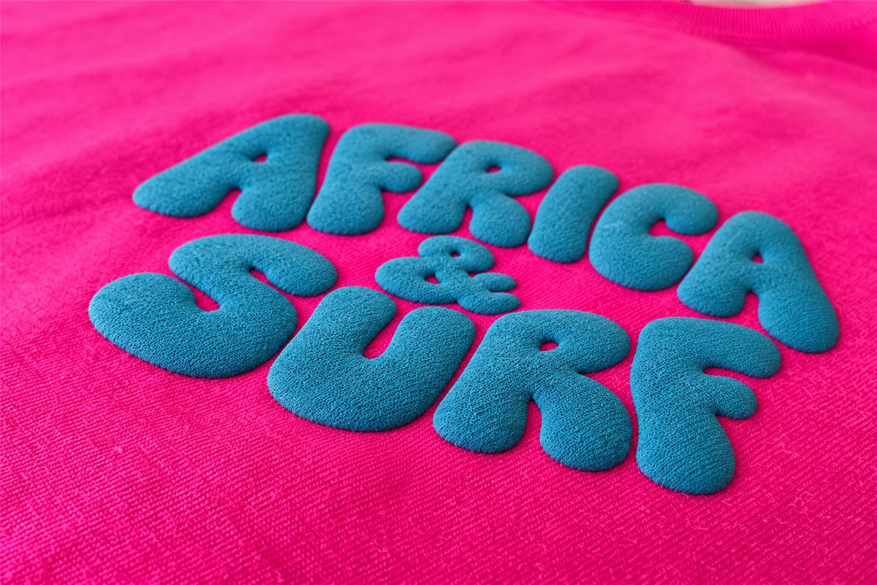 Raised teal puff print reading Africa & Surf on a pink tee