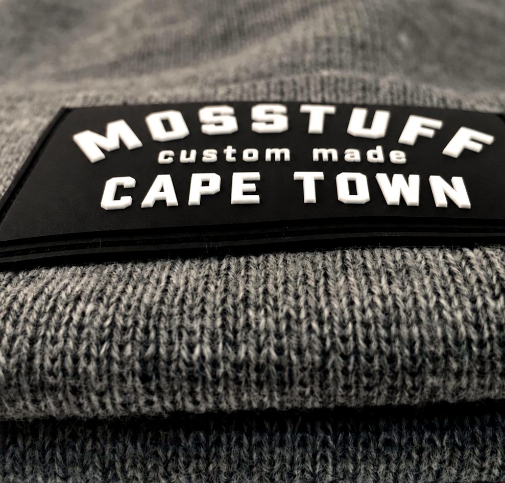 Black silicone rubber badge reading Mosstuff custom made Cape Town on a grey beanie